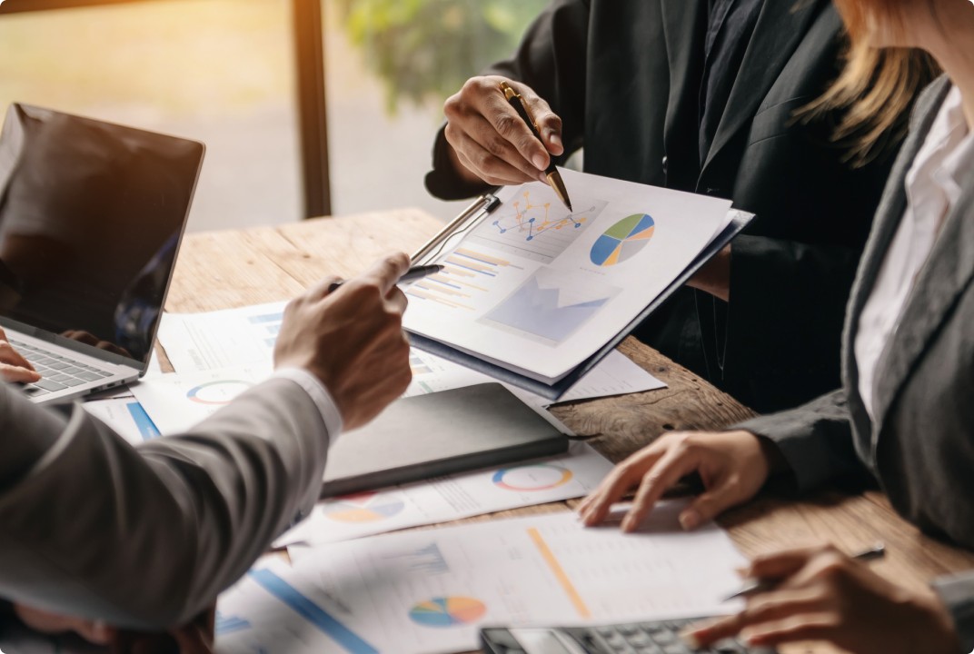 Três pessoas em um ambiente de escritório revisando gráficos financeiros em relatórios impressos, discutindo análises de mercado e dados de desempenho com gráficos de pizza e de barras.