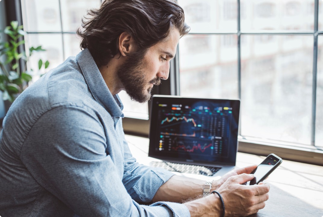Homem em um escritório analisando gráficos financeiros em um smartphone, com um laptop ao fundo exibindo gráficos de ações, sugerindo um ambiente de trabalho relacionado ao mercado financeiro ou investimentos.