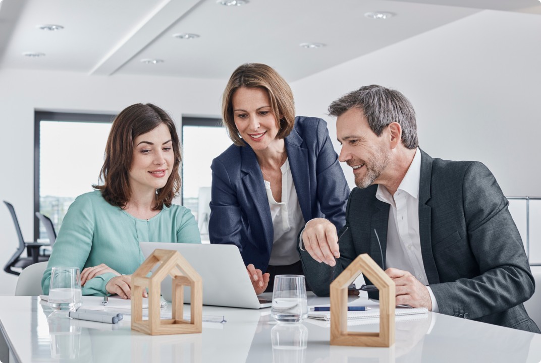 Três profissionais em um escritório moderno discutindo em frente a um laptop, com maquetes de casas sobre a mesa, representando uma reunião sobre investimentos imobiliários ou planejamento financeiro.